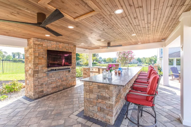 Outdoor patio with stone bar, red barstools, TV, ceiling fans, and garden view.