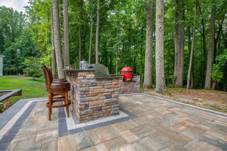 Outdoor patio with stone bar, two chairs, grill, and trees in the background.