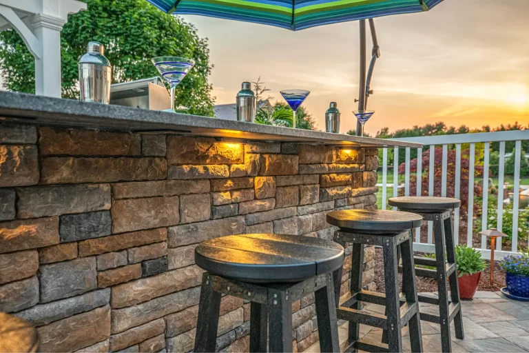 Outdoor stone bar with stools, martini glasses, and cocktail shakers under a striped umbrella at sunset.