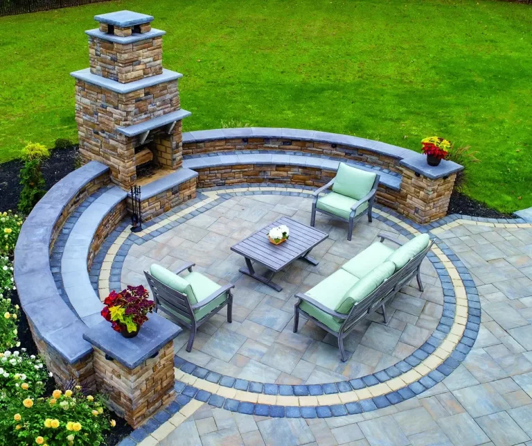 Stone patio with seating, a fireplace, and green lawn. Cushioned chairs and potted plants decorate the area.