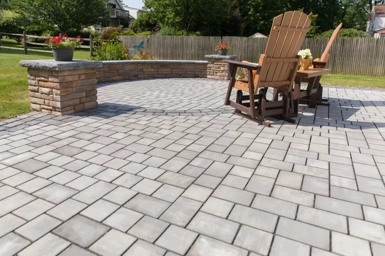 Two wooden chairs sit on a spacious stone patio with a low stone wall and potted flowers.