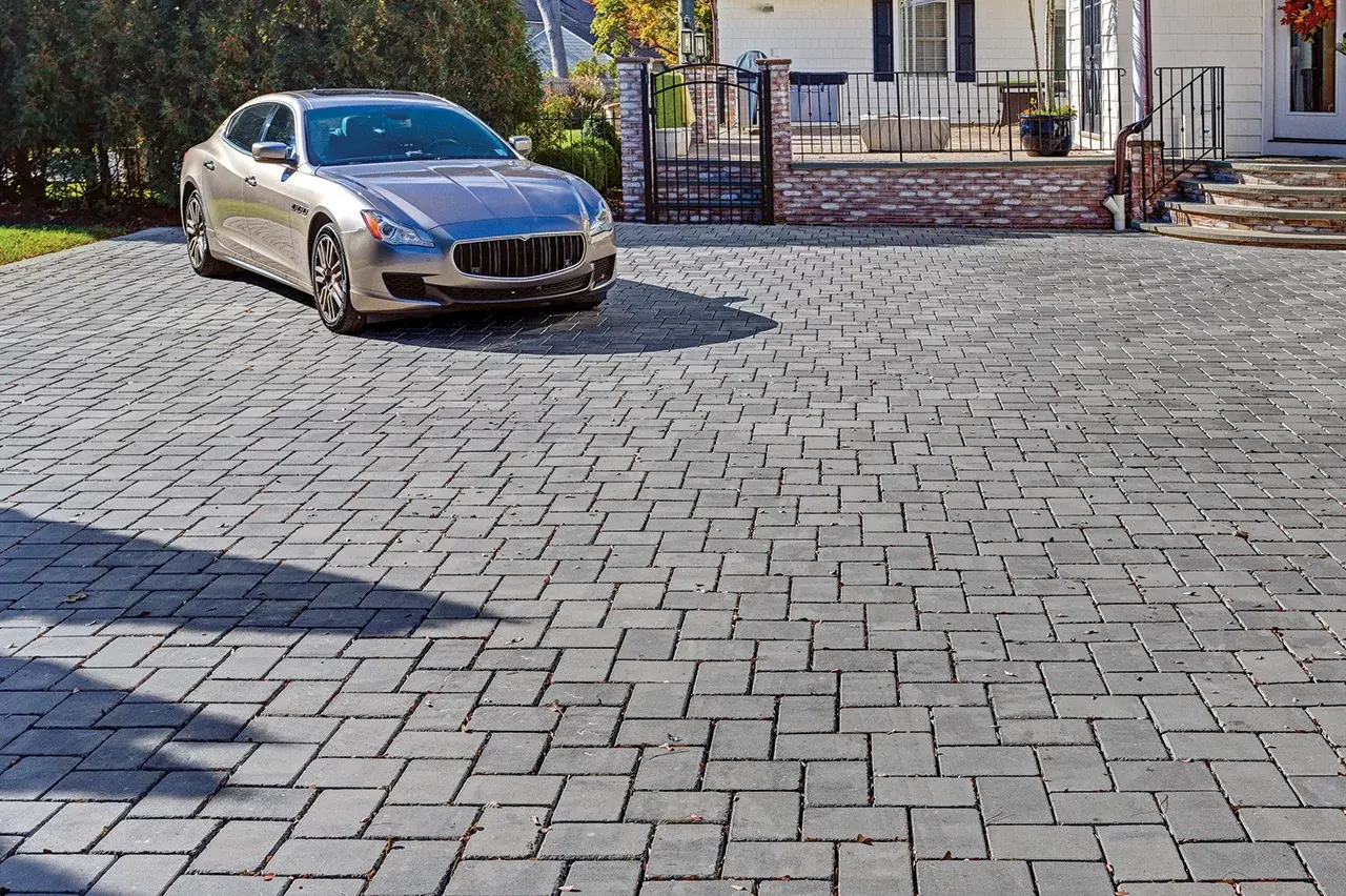A newly paved black asphalt driveway curves past a brick house with green lawns and landscaping, bordered by stone edging and surrounded by tall trees.