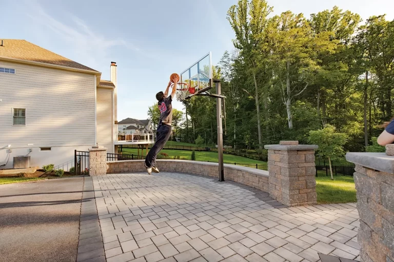 Person jumping to dunk a basketball on an outdoor hoop in a suburban backyard with trees and houses nearby.