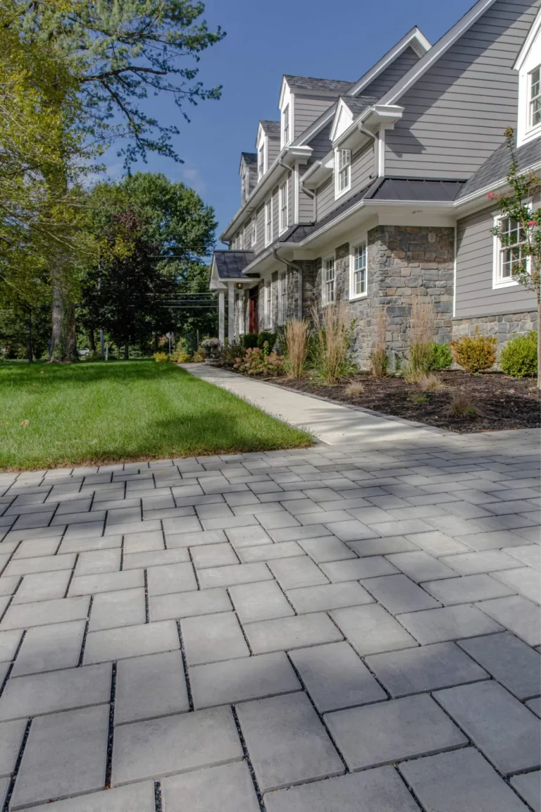 Modern house with gray siding, stone accents, and a large gray paver driveway, surrounded by grass and trees.