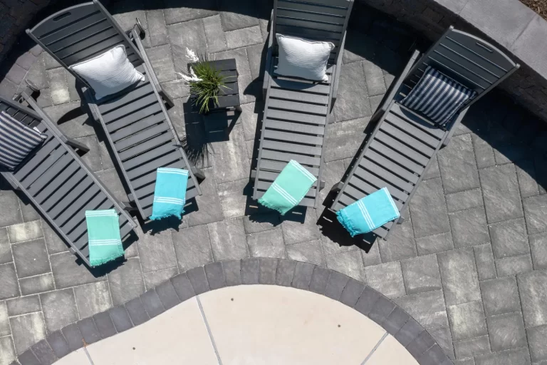 Four gray lounge chairs with towels and pillows are arranged beside a curved pool on a stone patio.