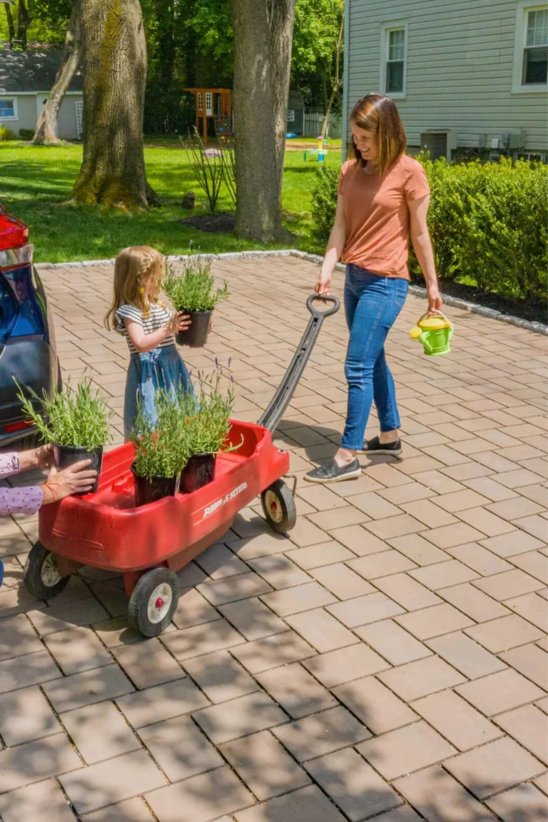 A woman pulls a red wagon with plants while a young girl holds a potted plant in a driveway.