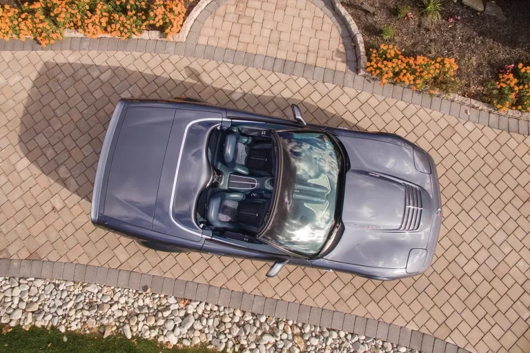 A silver convertible car on a stone driveway, surrounded by flowers and landscaping, viewed from above.