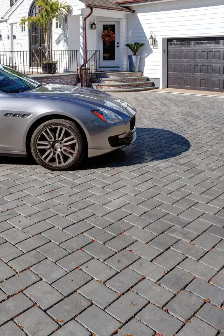 Silver car parked on a driveway with gray stone pavers in front of a white house and black garage door.