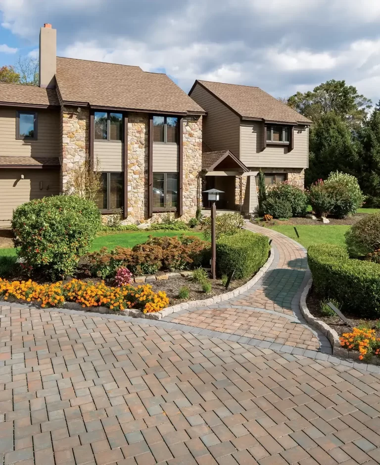 A stone pathway and driveway lead to a modern house with a garden and colorful flower beds.