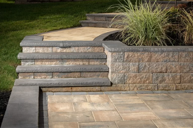 Stone steps and retaining wall beside a paved patio, with grass and ornamental grass plant above.