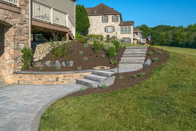 Stone steps with mulch and young plants lead up to a house in a landscaped backyard on a sunny day.