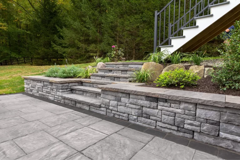 Stone patio with tiered steps, retaining wall, metal railing, and landscaped garden in a wooded backyard.