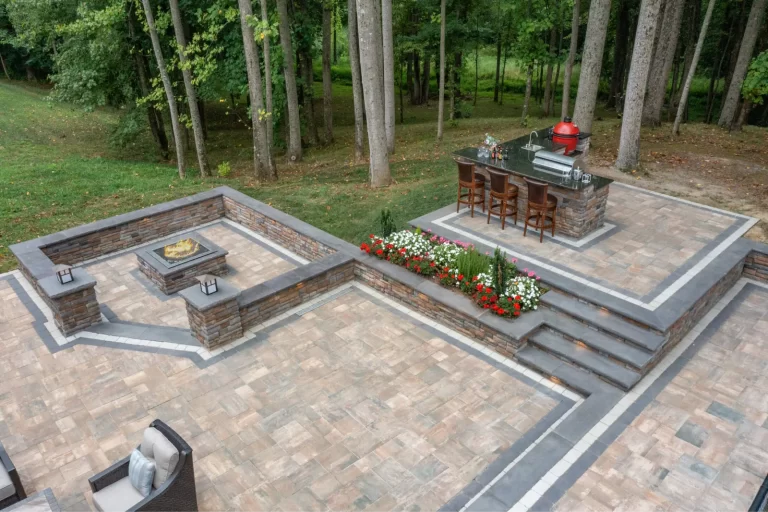 Stone patio with fire pit, outdoor kitchen, bar seating, and flower bed, surrounded by trees.