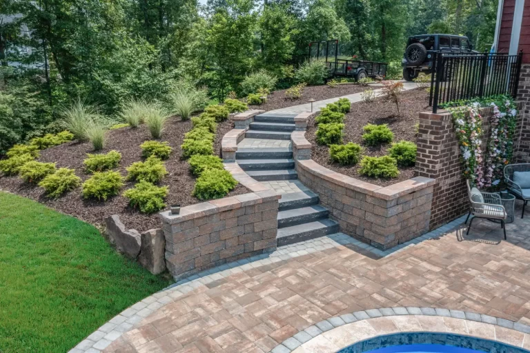 Curved stone steps with retaining walls and landscaping lead up from a patio and pool area to a wooded yard.
