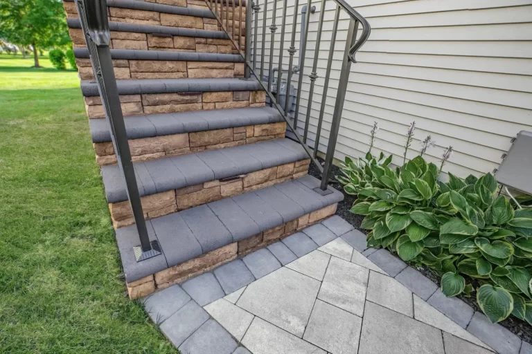 Stone steps with black railing leading to a house, bordered by green grass and leafy plants.