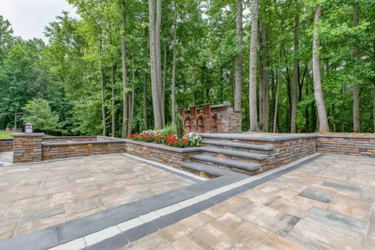 Spacious stone patio with raised flower bed, steps, and outdoor bar seating set among tall trees.