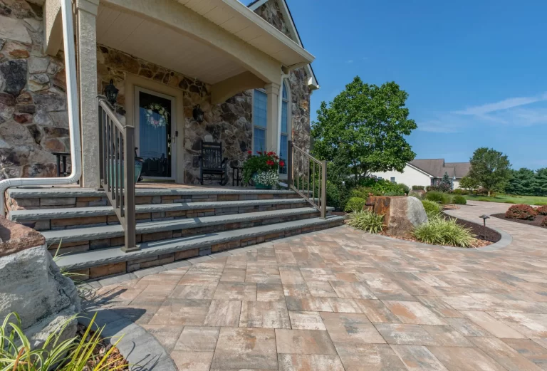 Stone steps lead to a porch with chairs; paved walkway, landscaping, and trees in front of a house.