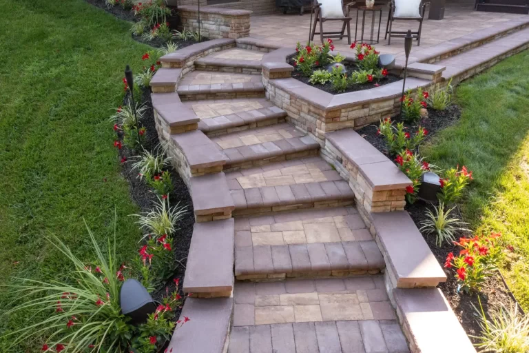 Stone steps with brick planters, surrounded by red flowers and greenery, leading to a patio with chairs.