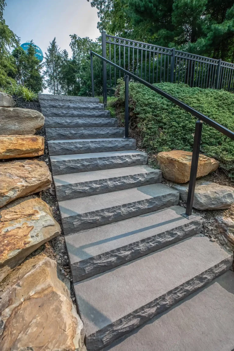 Stone steps with black railings, bordered by rocks and greenery, leading up a landscaped slope.
