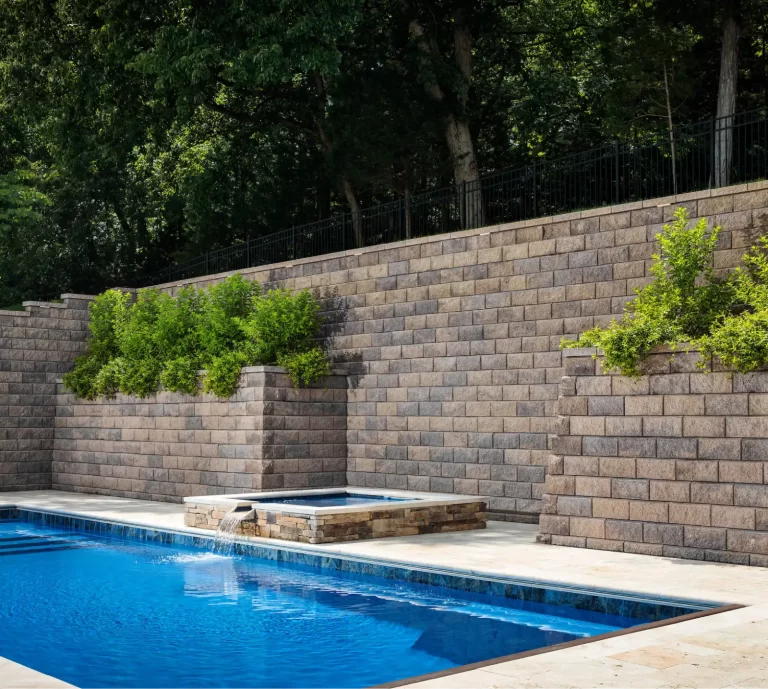 In-ground pool and hot tub with waterfall, bordered by a stone retaining wall and green shrubs.