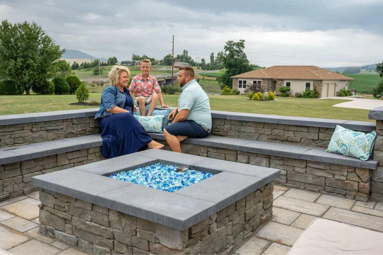 Three people sit and talk around a modern outdoor fire pit with blue glass, surrounded by stone benches.