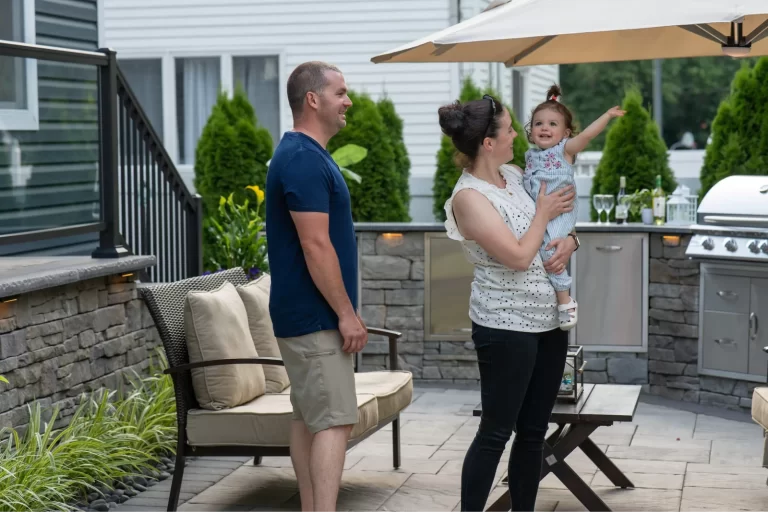 A woman holding a smiling toddler points up while a man stands beside them on a patio.