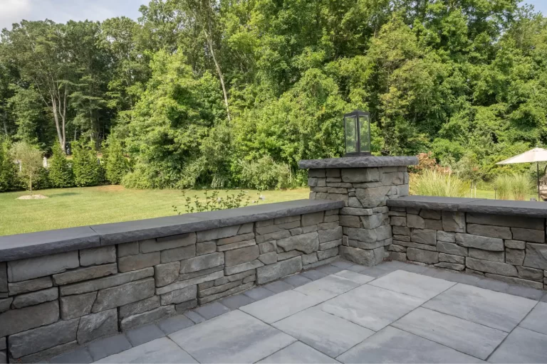 Stone patio with a short stone wall, overlooking a grassy yard and dense green trees in the background.