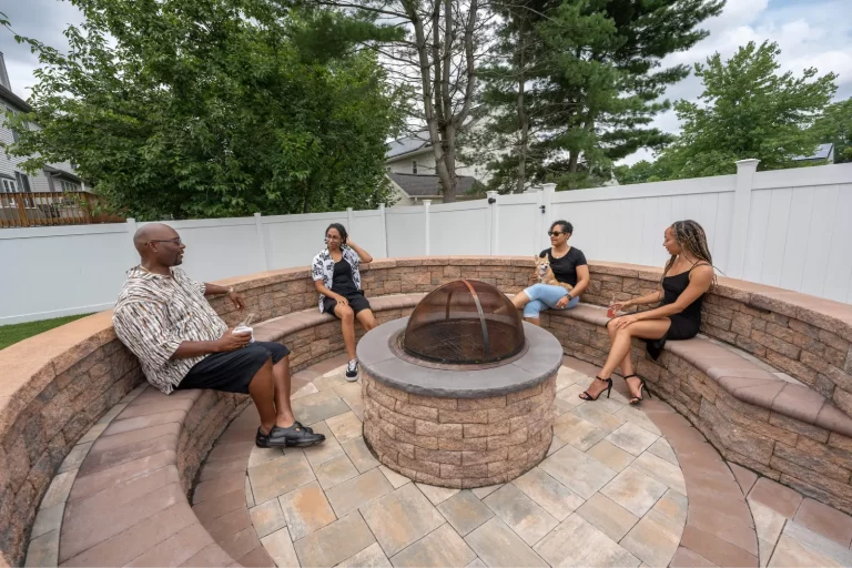 Four people sit and chat around a circular stone fire pit in a backyard patio area.