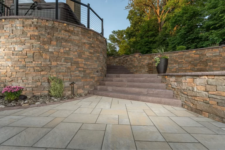 Stone staircase and patio with a curved stone wall, potted plants, and greenery in the background.