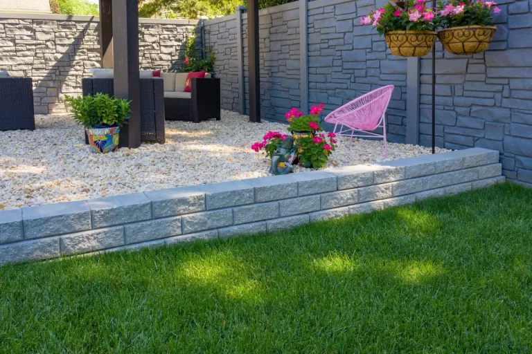 Modern backyard with stone wall, green grass, potted flowers, and outdoor seating on white gravel.