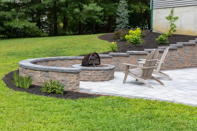 Two wooden chairs on a stone patio with a fire pit, surrounded by grass, plants, and a retaining wall.