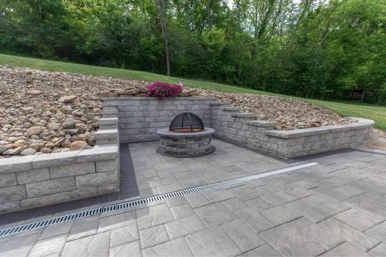 Stone patio with a circular fire pit, retaining walls, and a flower planter, surrounded by trees and rocks.