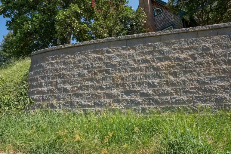 A stone retaining wall with grass in front and trees and a house partially visible behind it.
