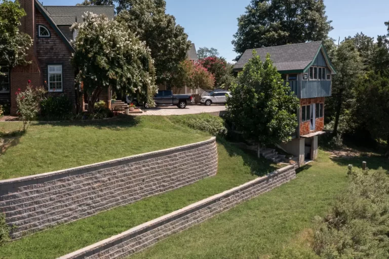 Two-tiered retaining wall supports a grassy slope by houses and parked cars on a sunny day.