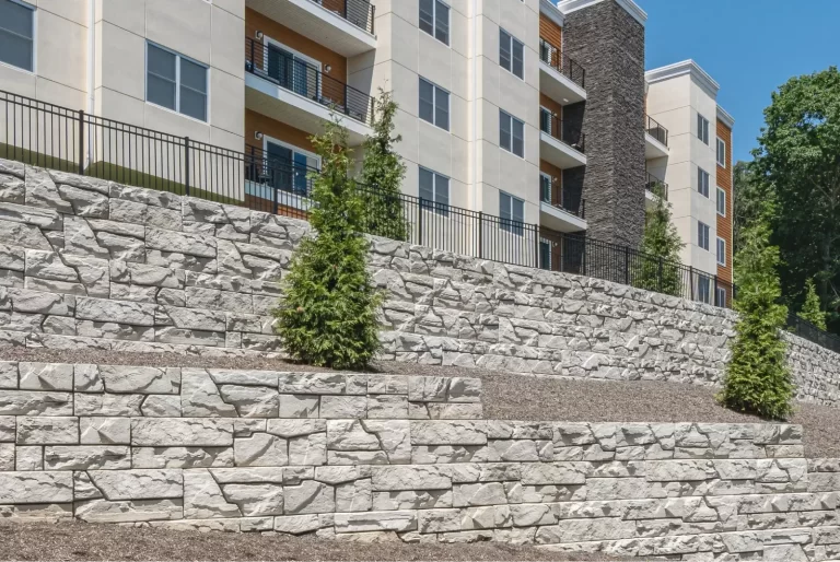 Terraced stone retaining walls with young trees in front of a modern apartment building on a sunny day.