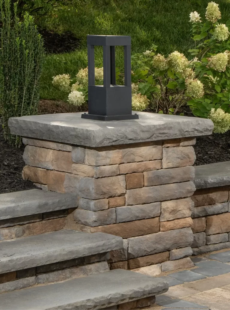 Stone pillar with a square black outdoor lantern, surrounded by greenery and blooming white flowers.