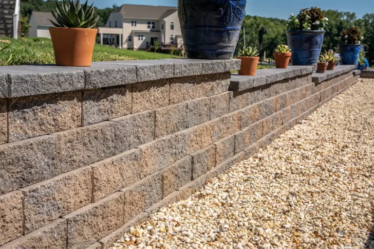 Stone retaining wall with potted plants on top and a gravel path beside it in a suburban yard.