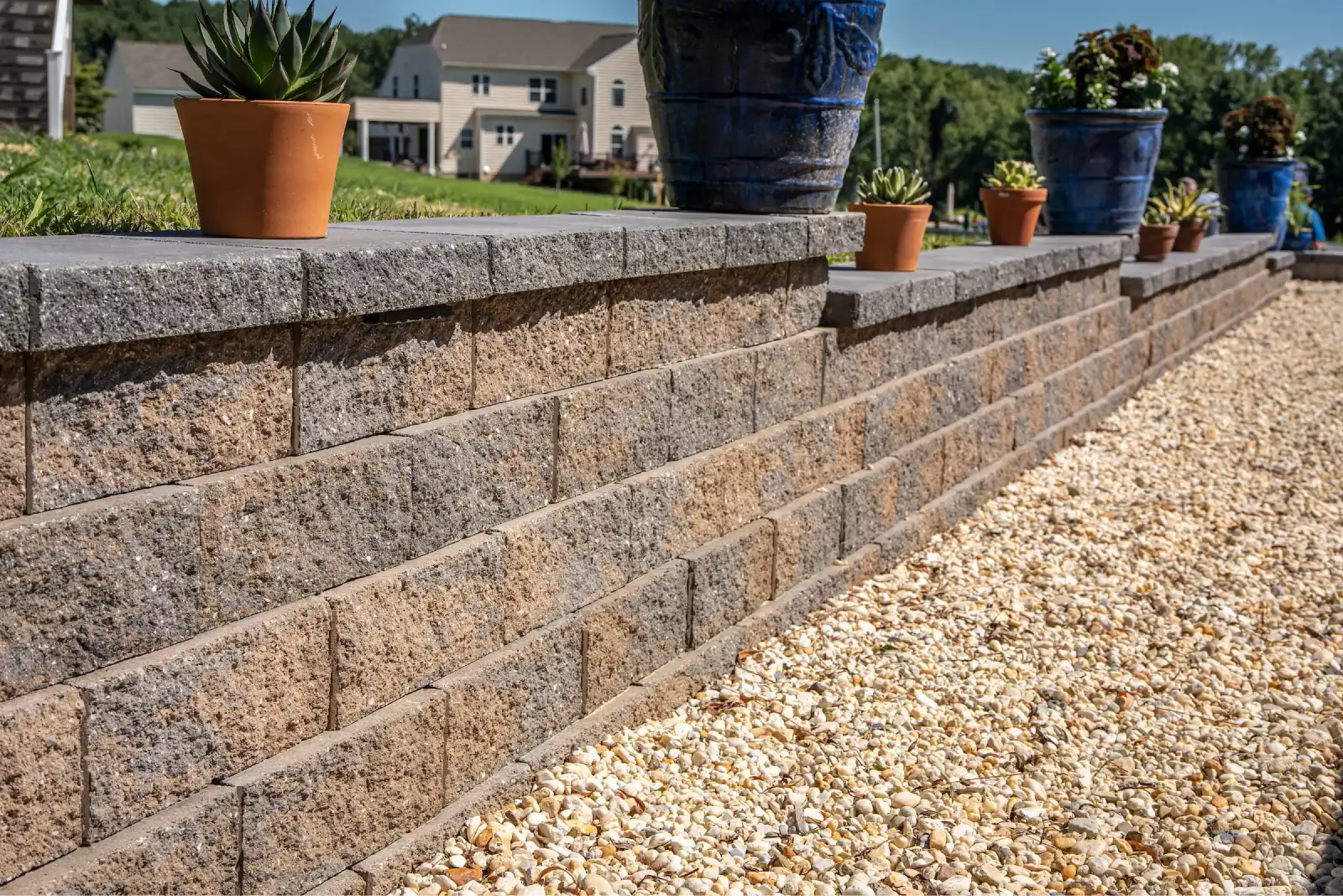 Stone retaining wall with potted plants on top and a gravel path beside it in a suburban yard.