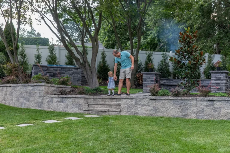 Man helping a small child walk down stone steps in a landscaped backyard with trees and greenery.