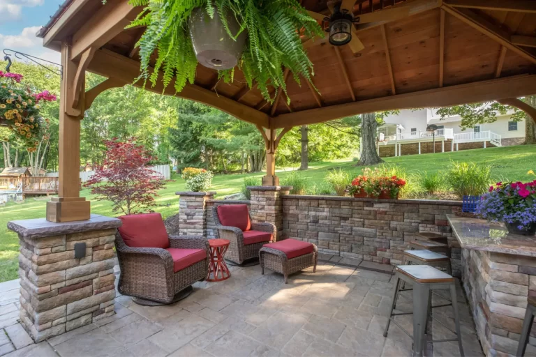 Covered patio with wicker chairs, red cushions, stone walls, and lush plants in a green backyard.