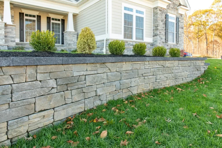 Stone retaining wall with neatly trimmed shrubs in front of a house and green lawn with scattered leaves.