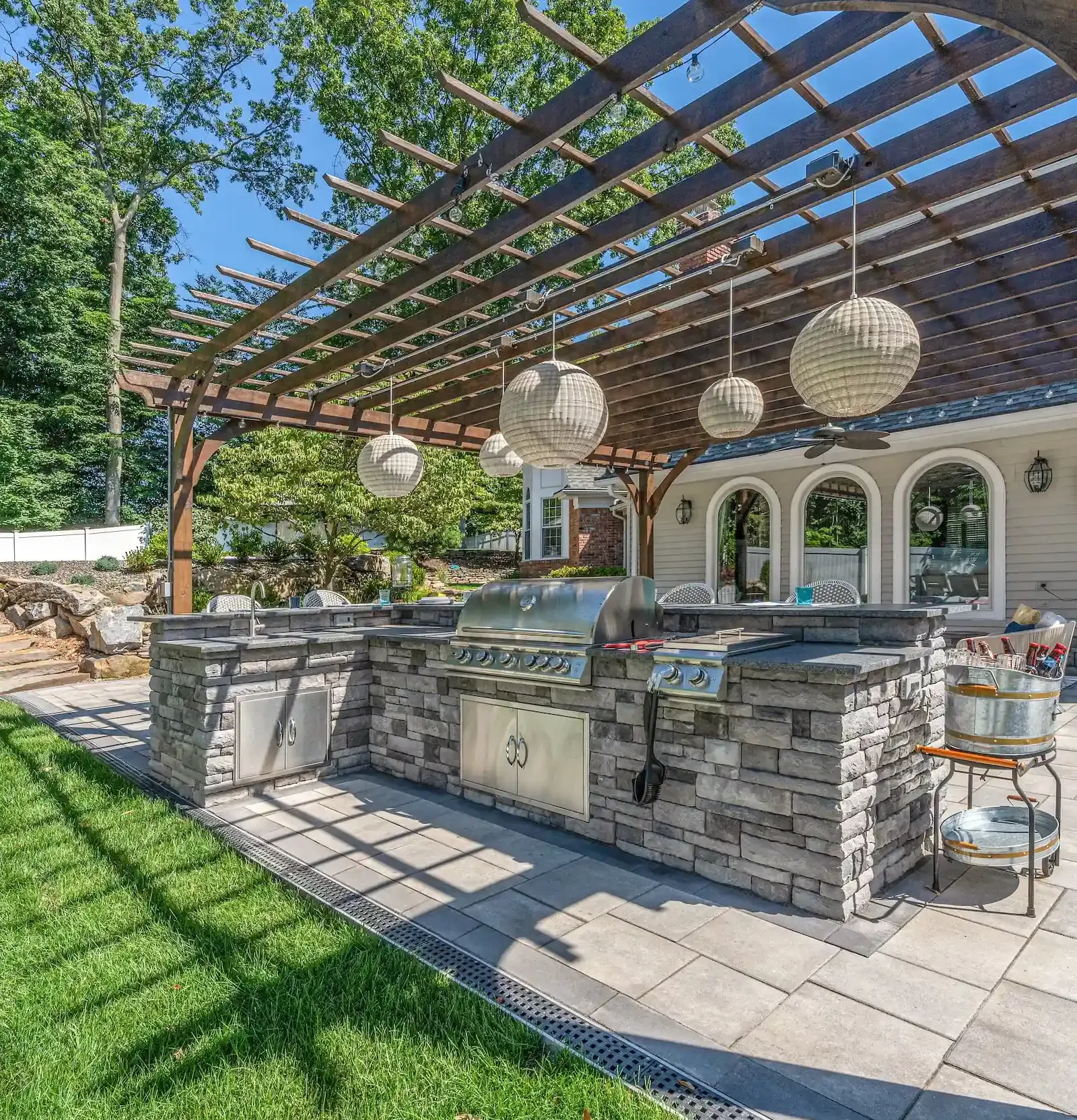 Outdoor kitchen with stone counters, grills, hanging lanterns, and a wooden pergola over a patio area.