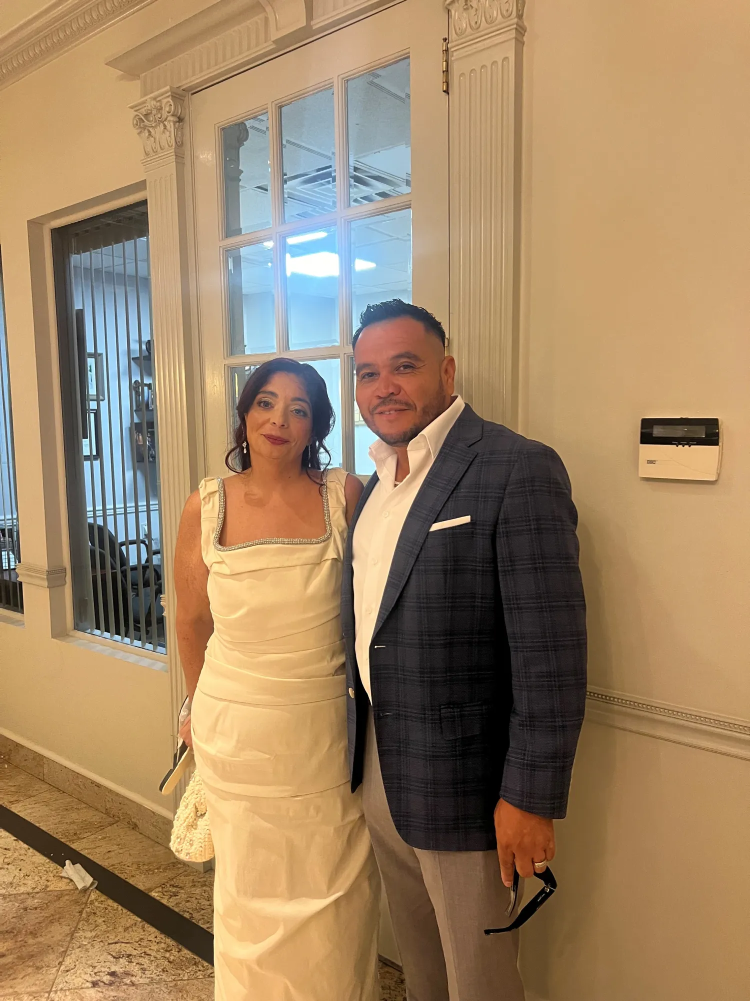 A woman in a white dress and a man in a plaid blazer pose together indoors by a glass door.