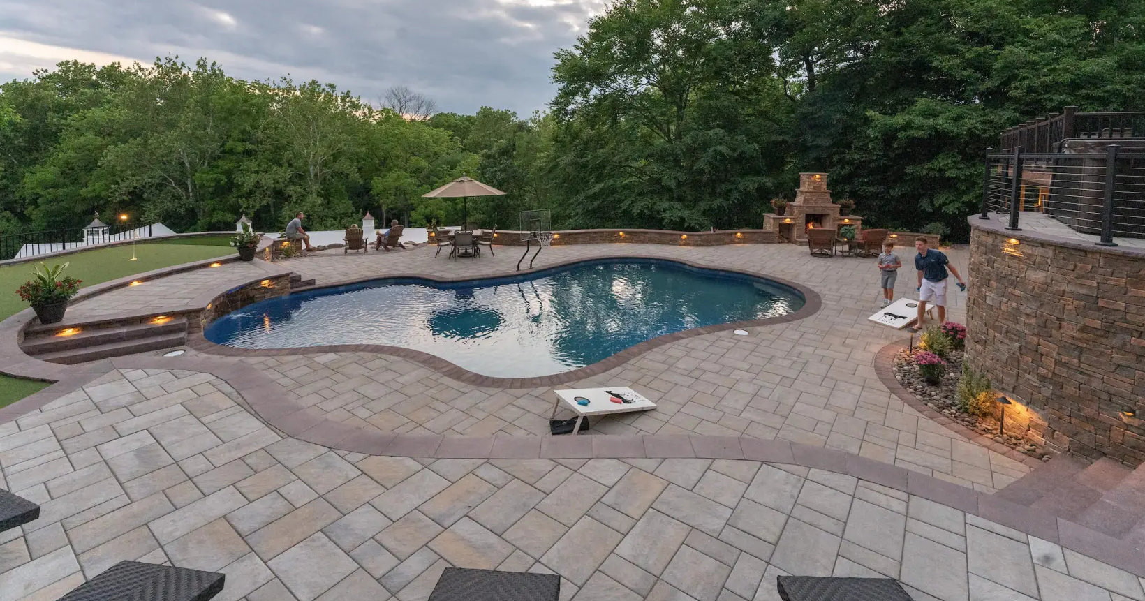 Backyard pool with patio, cornhole boards, seating, people relaxing, and trees in the background at dusk.